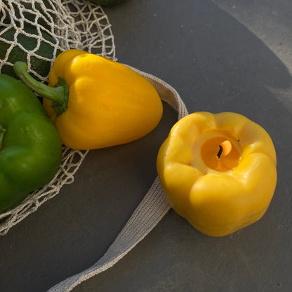 Bell Pepper Candle - Yellow next to fresh green peppers in a net bag, perfect artisan decor piece for your home.