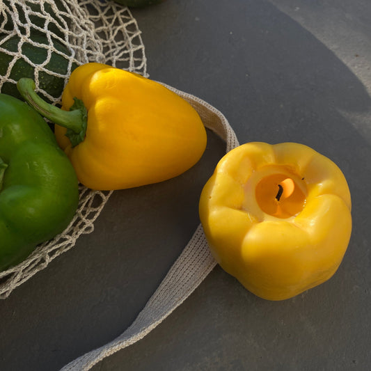 Bell Pepper Candle - Yellow next to fresh green peppers in a net bag, perfect artisan decor piece for your home.