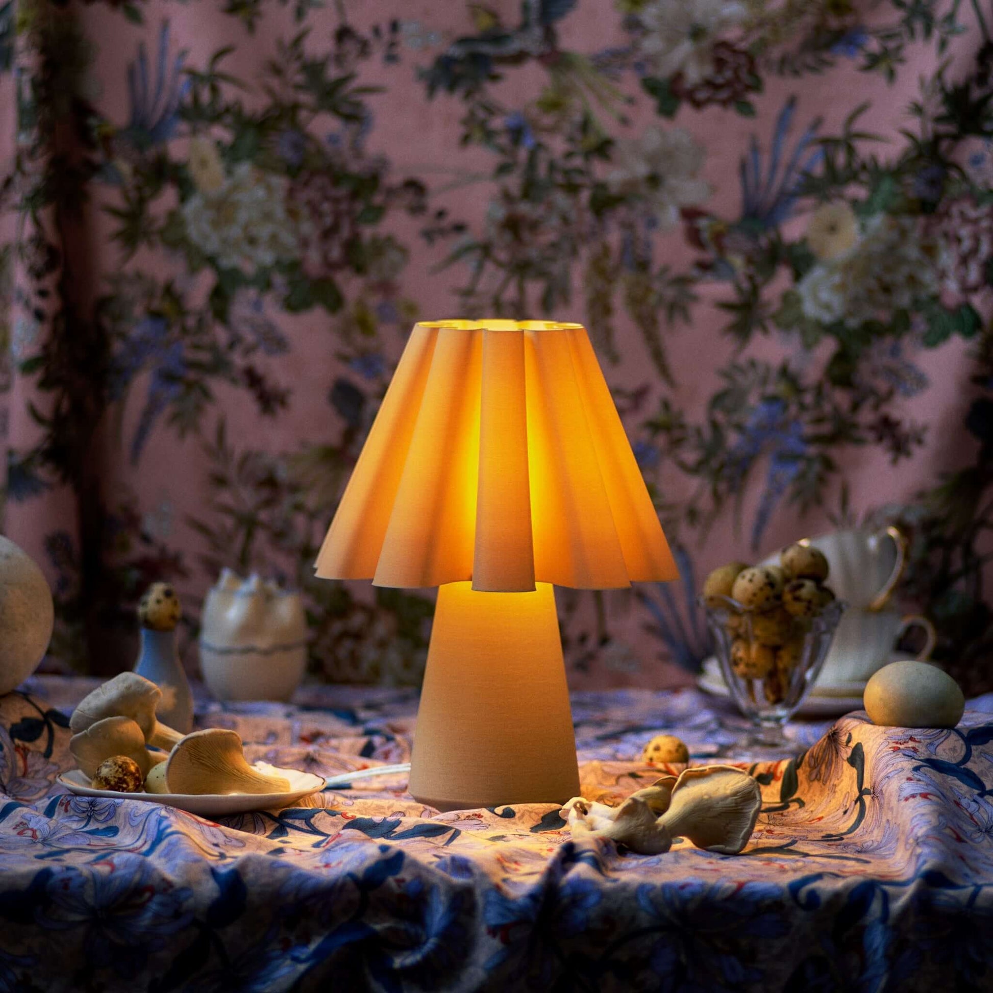 Table lamp with a warm glow on a decorative tablecloth against a floral wallpapered wall.